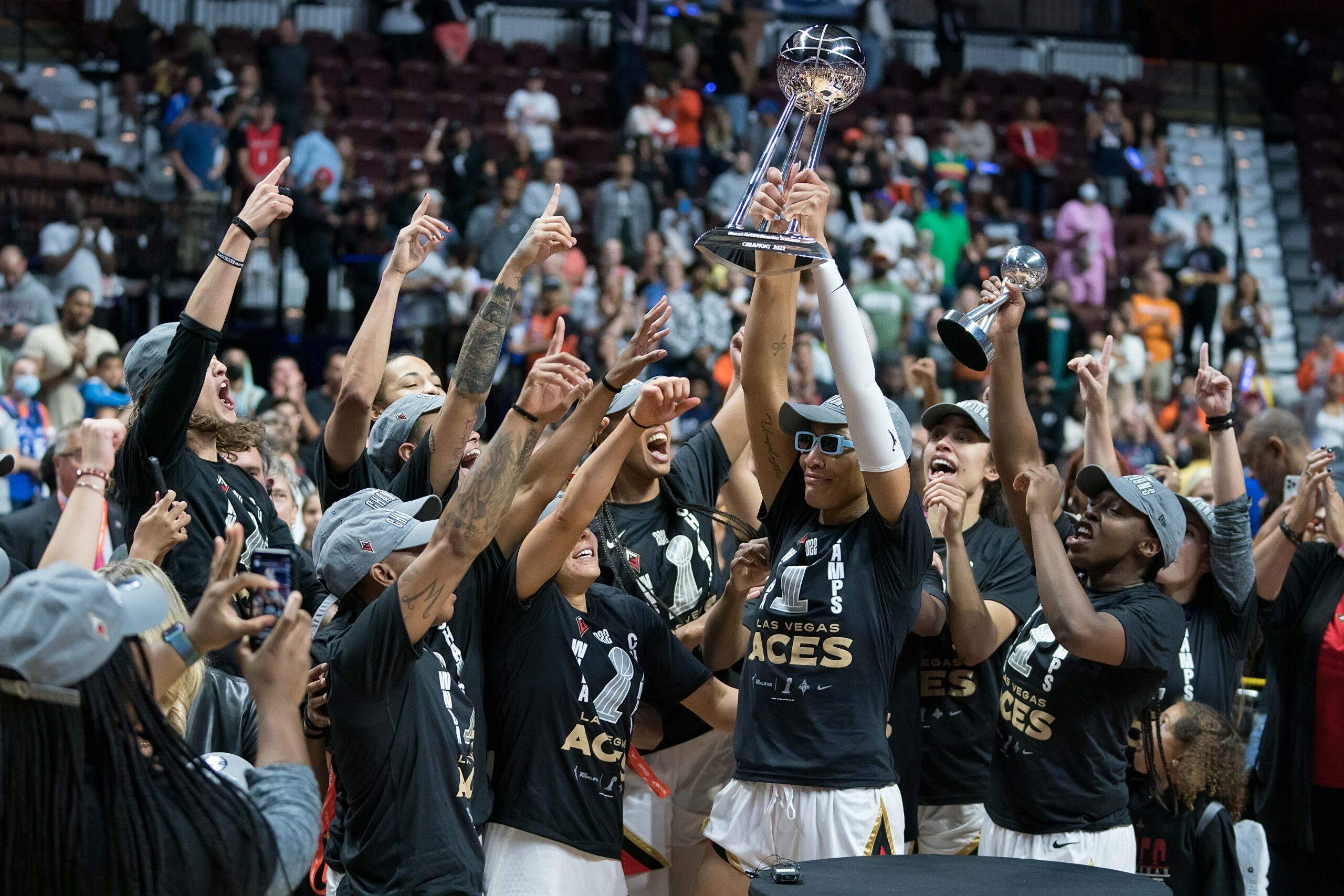 Las Vegas Aces center A'ja raises the 2022 WNBA championship trophy. The rest of the Aces team celebrate around her, with their arms in the air.