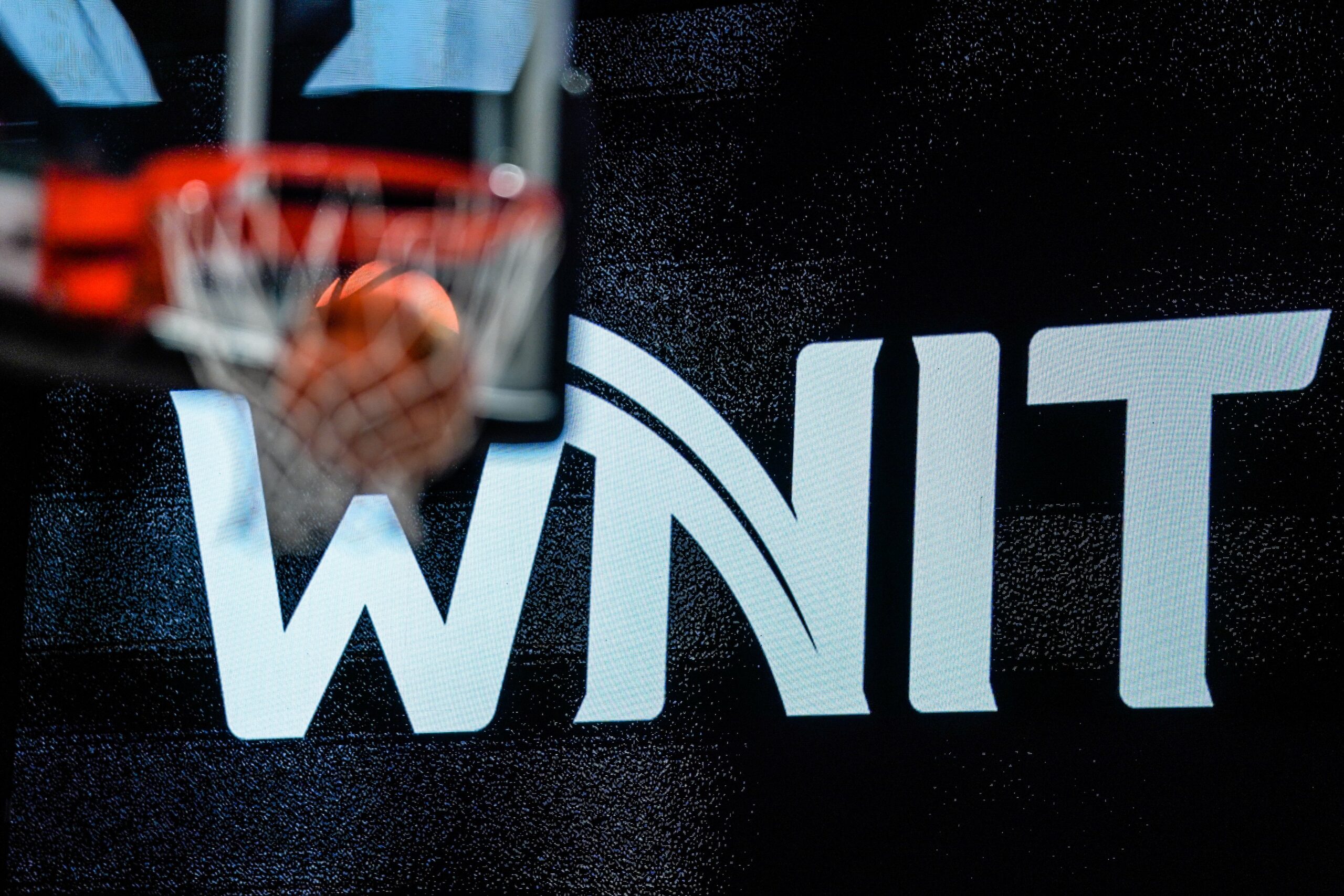The WNIT logo with a blurred out basketball hoop in the foreground. A ball goes through the hoop, which is placed just over the "W" in WNIT.