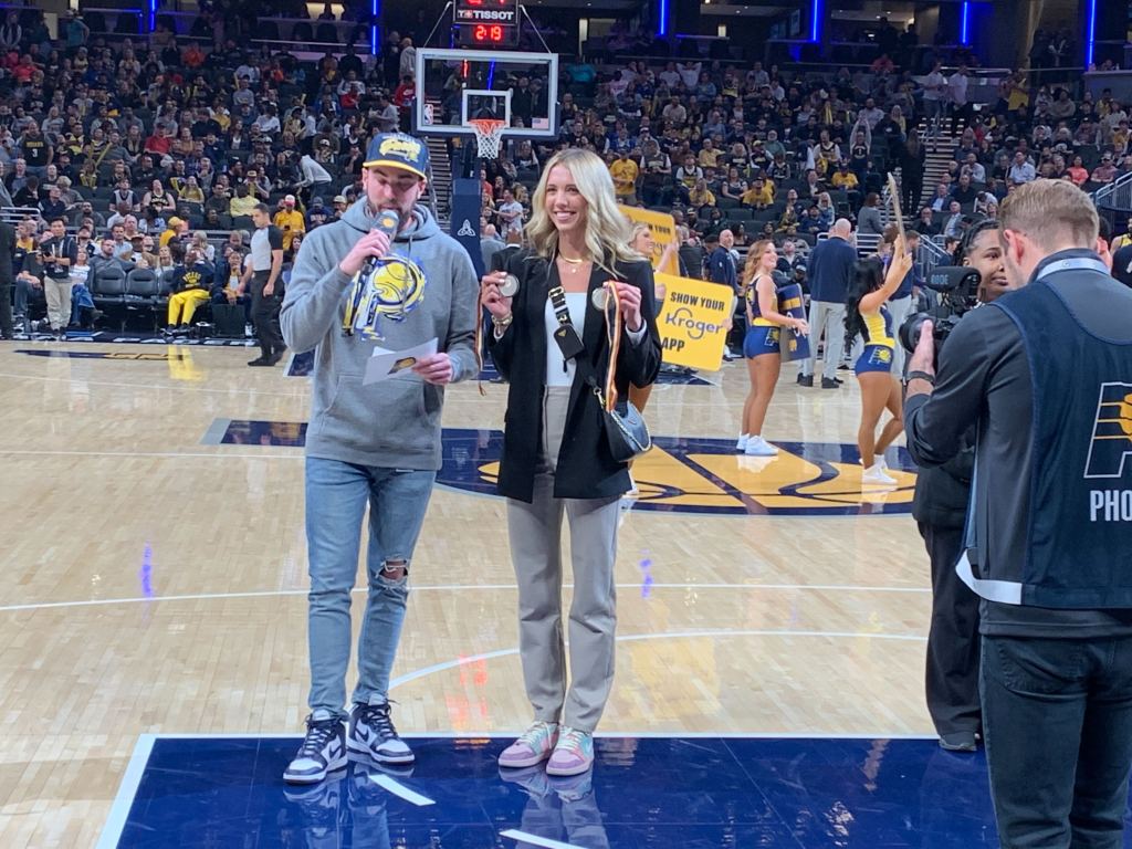 Lexie Hull poses with her silver medal from the 2022 FIBA U23 3x3 World Cup. A host of the recognition ceremony stands to her left, reading off of a notecard into a microphone.