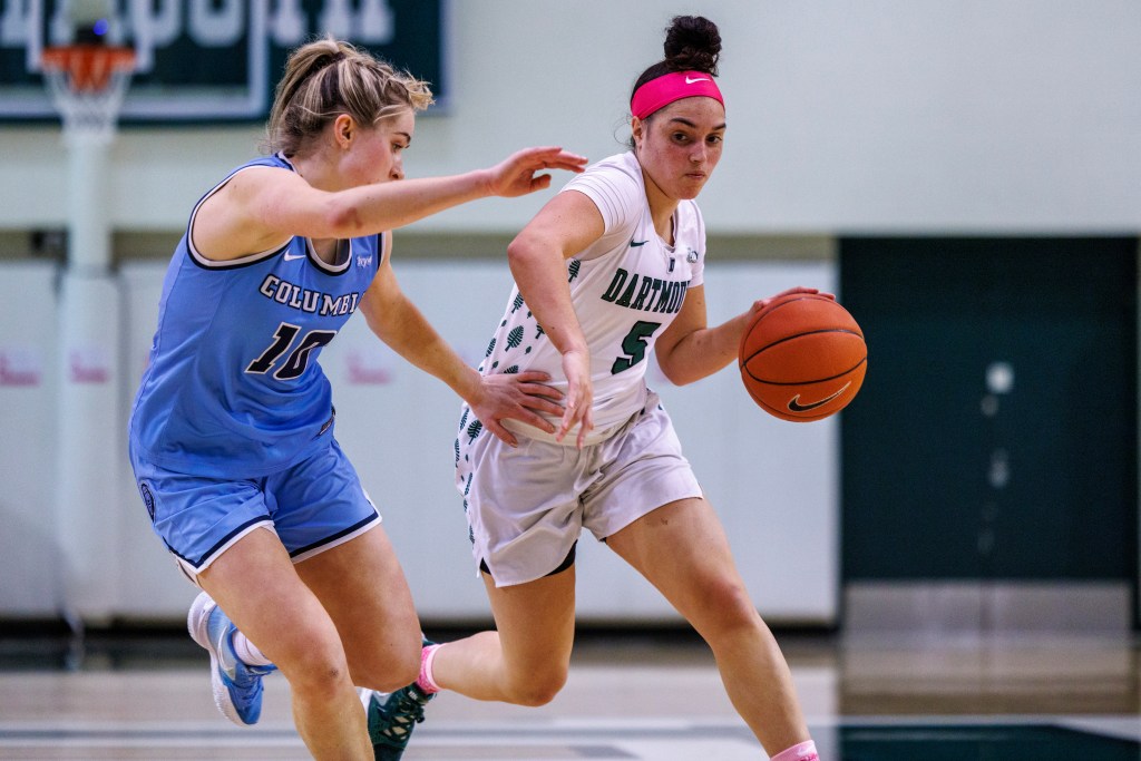 Dartmouth guard Mia Curtis, wearing a white jersey and bright pink headband, dribbles as Columbia guard Kitty Henderson tries to stay in front.