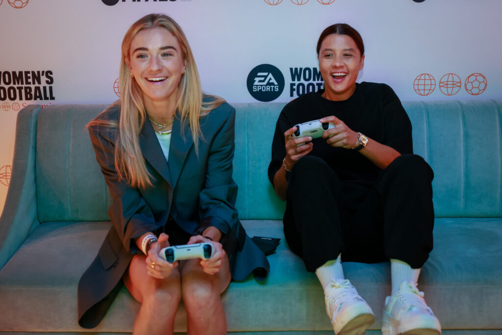 Photo of Kristie Mewis and Sam Kerr sitting on a couch and holding PlayStation controllers.