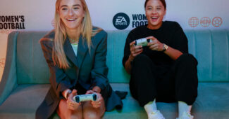 Photo of Kristie Mewis and Sam Kerr sitting on a couch and holding PlayStation controllers.