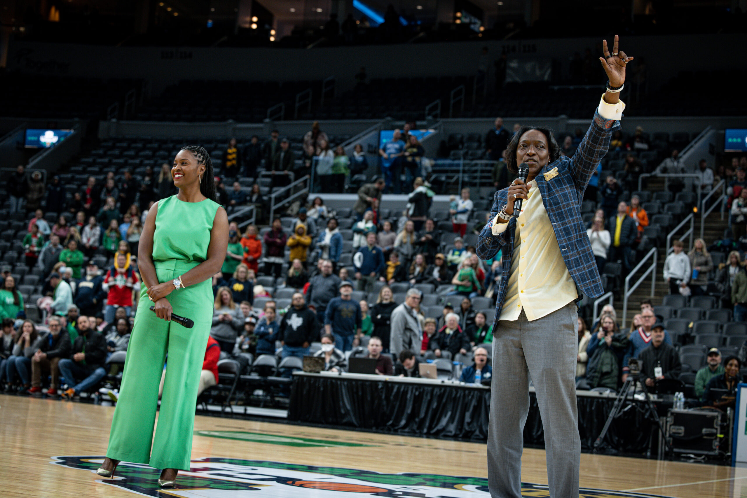 California head coach Charmin Smith raises her left hand in the air as she addresses the crowd, holding a microphone in her left hand. Notre Dame head coach Niele Ivey stands to her right, looking out at the crowd.