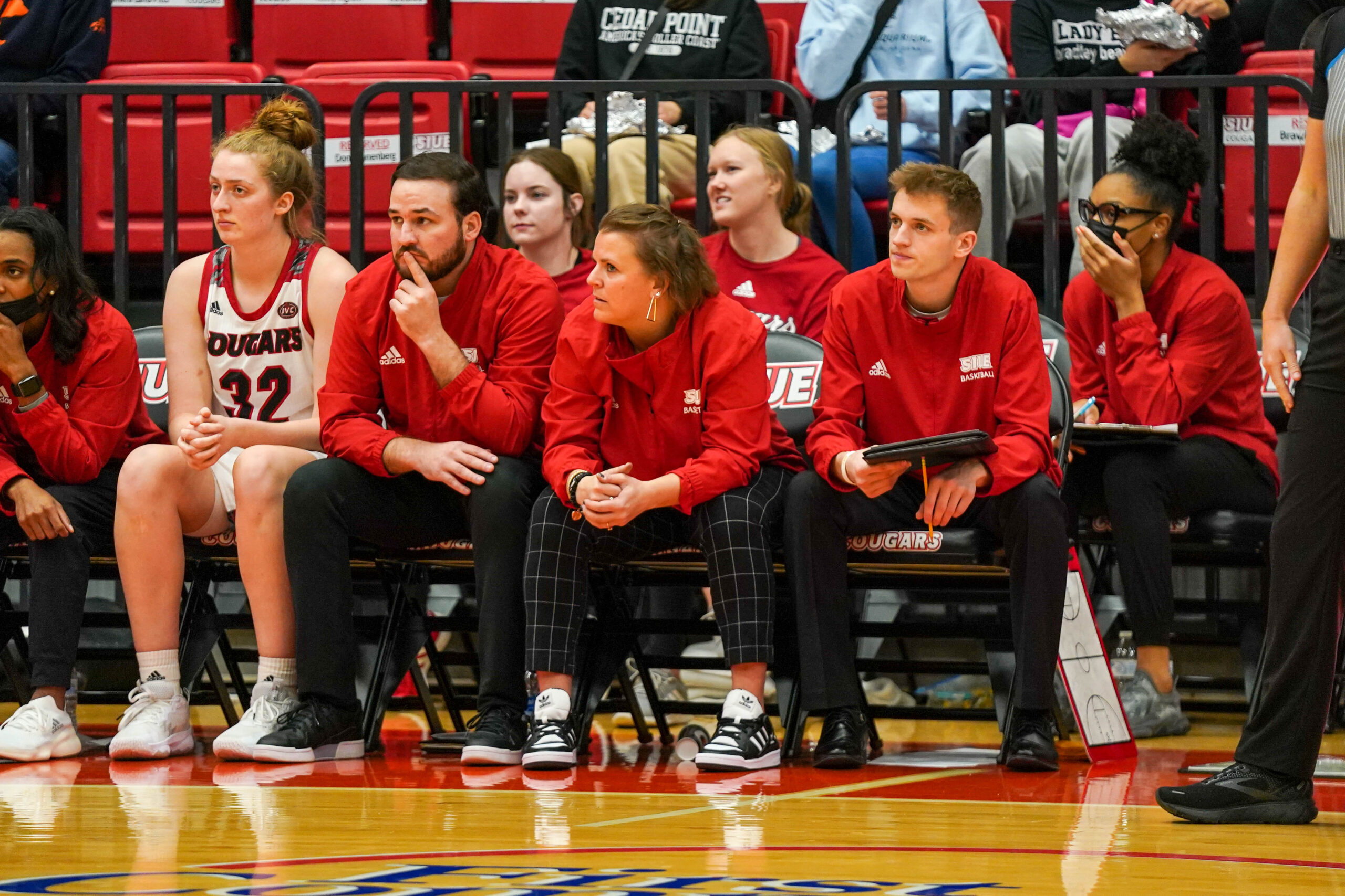 Players and coaches sit on the SIUE bench during a game against Xavier. Head coach Samantha Quigley Smith is looking downcourt, while assistant coach Bradley Bruno puts his left index finger to his chin.