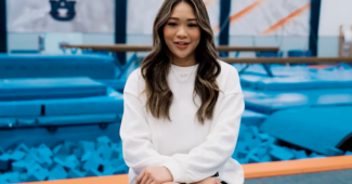 Gymnast Suni Lee sits with her hair down, wearing a white sweater with hands on her lap. She sits in front of the Auburn gymnastics gym.