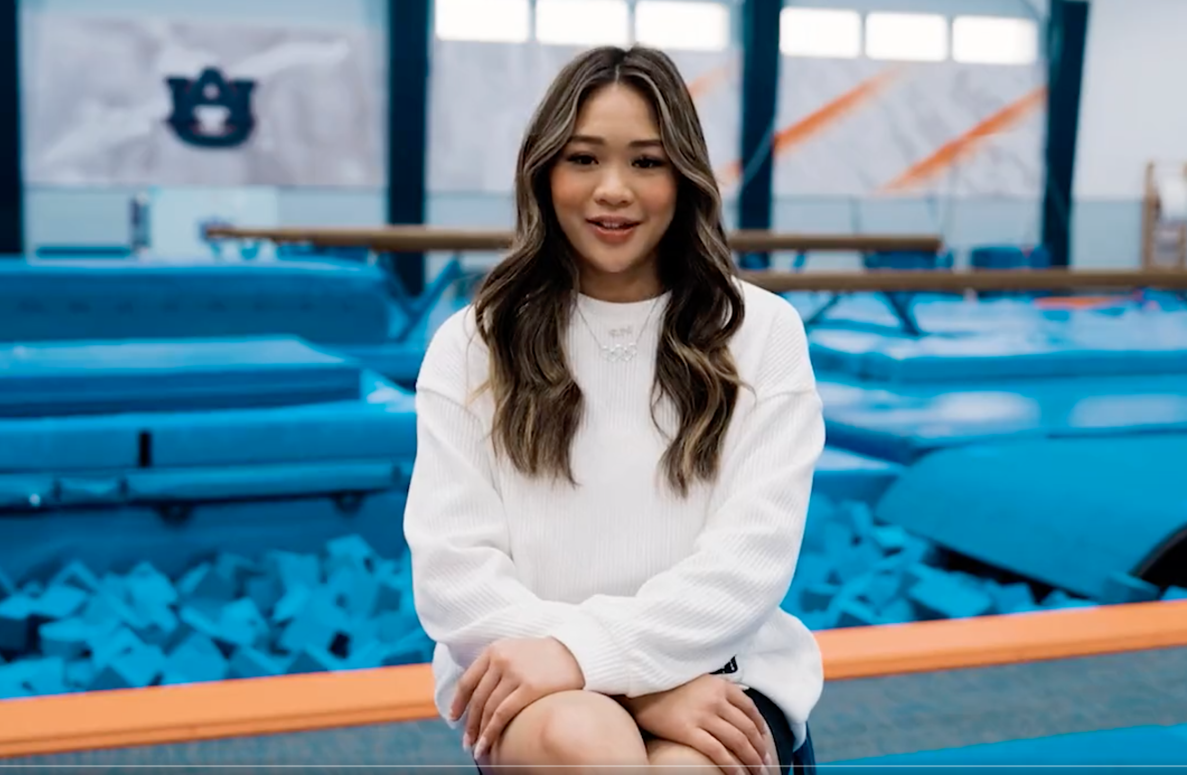 Gymnast Suni Lee sits with her hair down, wearing a white sweater with hands on her lap. She sits in front of the Auburn gymnastics gym.