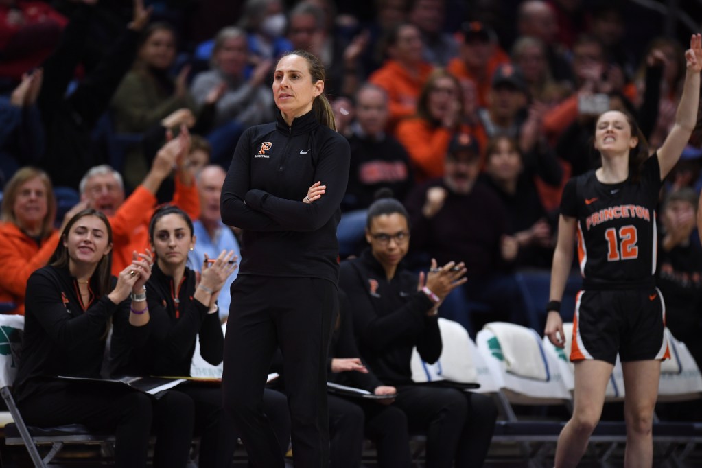 Princeton head coach Carla Berube stands with her arms crossed as assistant coaches clap behind her and a player on the bench stands to celebrate.