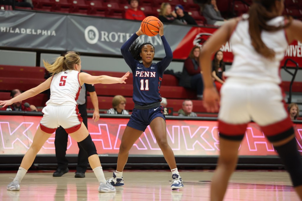 Penn guard Simone Sawyer holds the ball above her head with two hands and scans the court, with a defender in front of her.