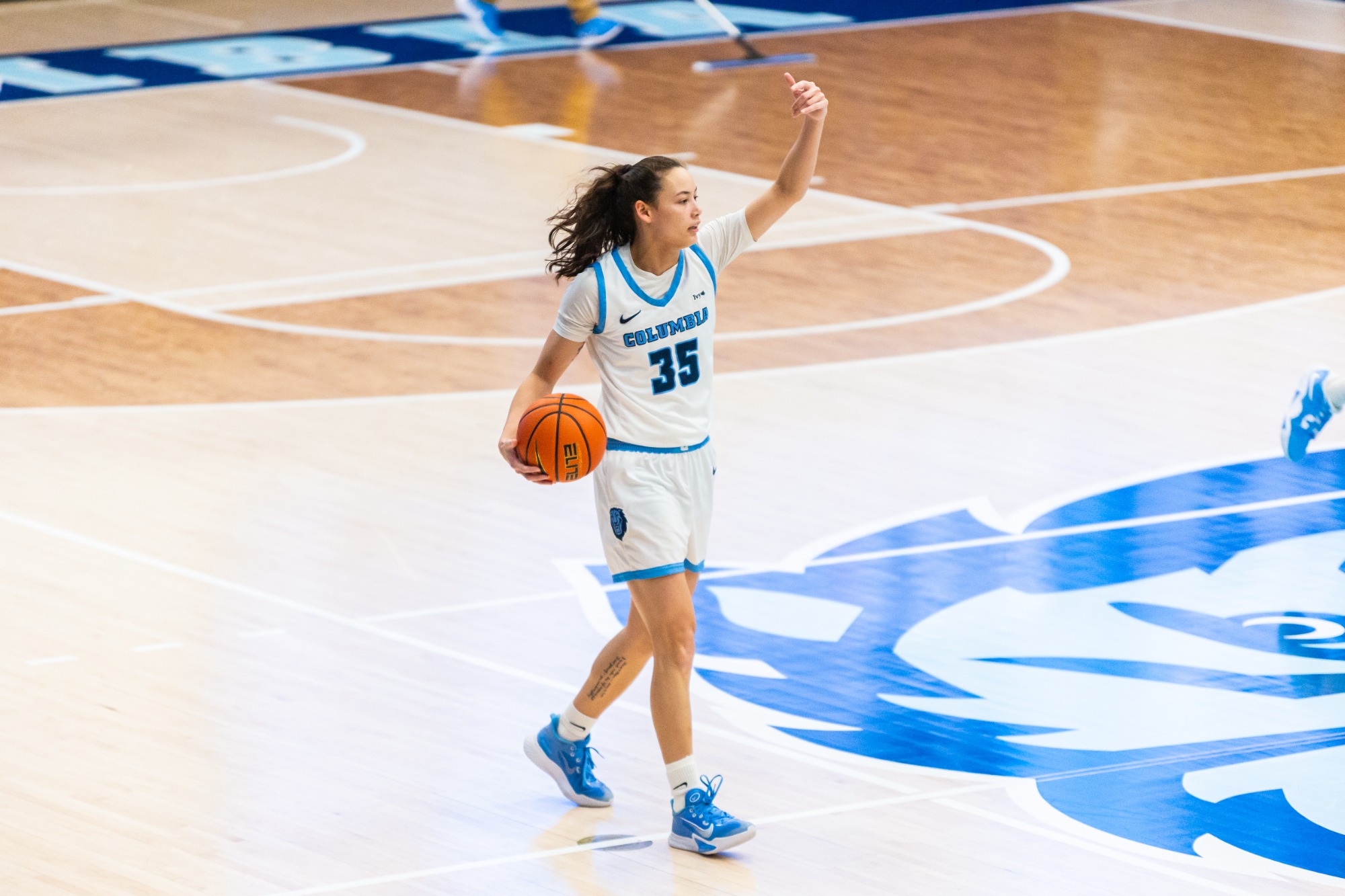 Abbey Hsu dribbles the ball across halfcourt with her right hand while gesturing to her teammates with her left hand. The large blue Columbia Lions logo at center court is visible just to Hsu's left.