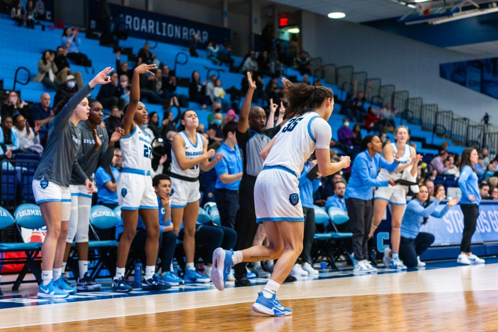 Columbia guard Abbey Hsu pumps her right fist as she turns to run back on defense after making a 3-pointer. Her teammates on the bench are visible behind her cheering and making the "3-point" sign with their hands.