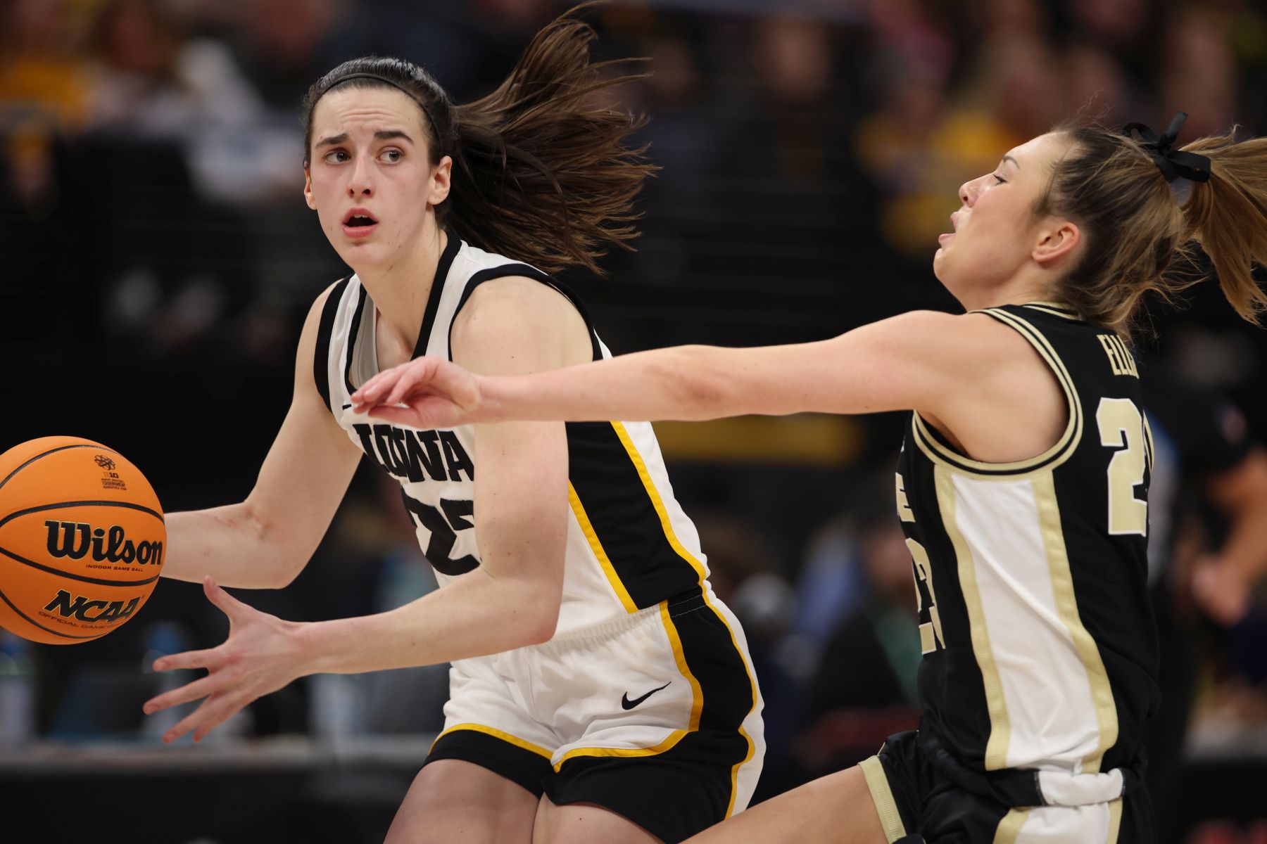 Iowa guard Caitlin Clark looks up across the court as she prepares to secure the basketball with two hands. A Purdue defender attempts to keep up with her.