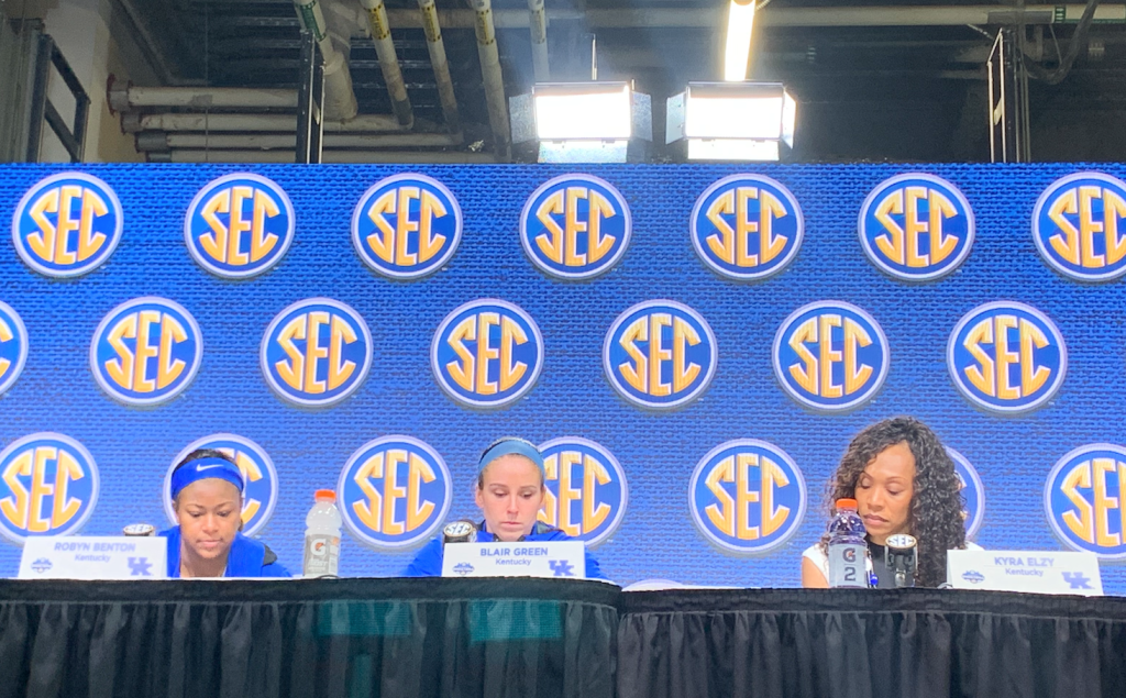 Two Kentucky players and head coach Kyra Elzy sit at a table with the SEC logo on a backdrop behind them. All look down at the table.