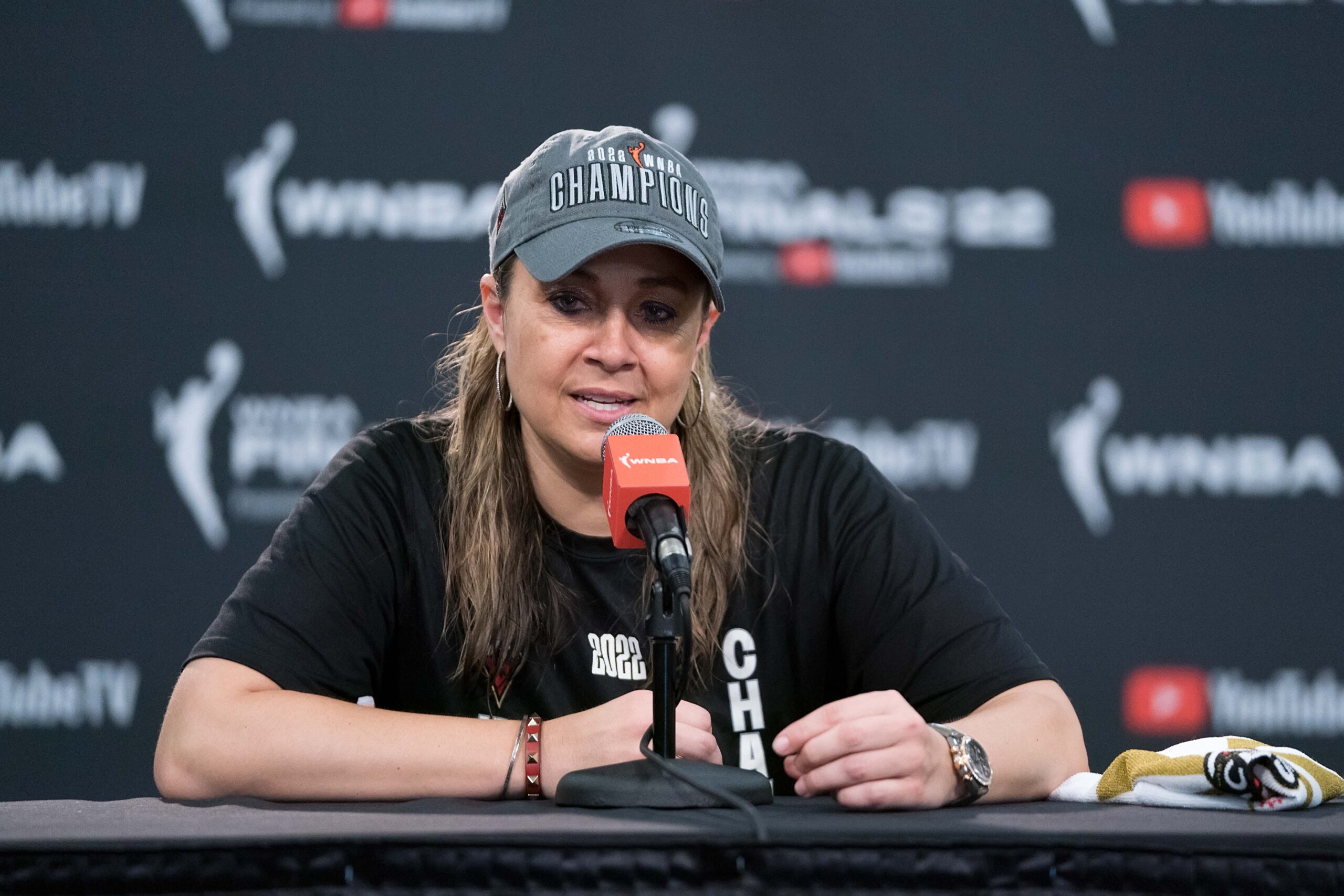 Wearing a baseball cap commemorating the Las Vegas Aces' 2022 WNBA championship, head coach Becky Hammon sits at the podium and answers questions from reporters.