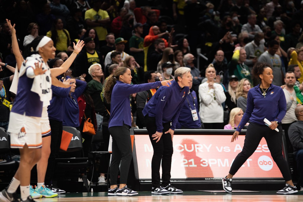 Phoenix Mercury assistant coach Cinnamon Lister points downcourt with her right arm. The Phoenix bench reacts to the play, and head coach Vanessa Nygaard watches with her hands on her thighs.