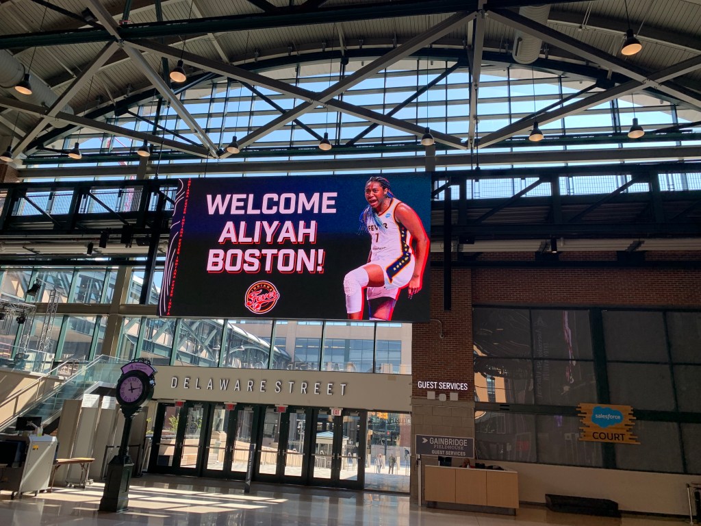 A sign above the Delaware Street entrance to Gainbridge Fieldhouse shows No. 1 overall WNBA Draft pick Aliyah Boston celebrating in an Indiana Fever uniform. The sign reads, "Welcome Aliyah Boston!" in all caps with a Fever logo below the text.