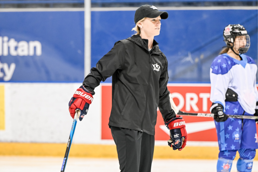 Kacey Bellamy skates at practice at the PHF All-Star Game. She is wearing a black PHF-branded jacket and hat and red hockey gloves.