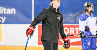 Kacey Bellamy skates at practice at the PHF All-Star Game. She is wearing a black PHF-branded jacket and hat and red hockey gloves.