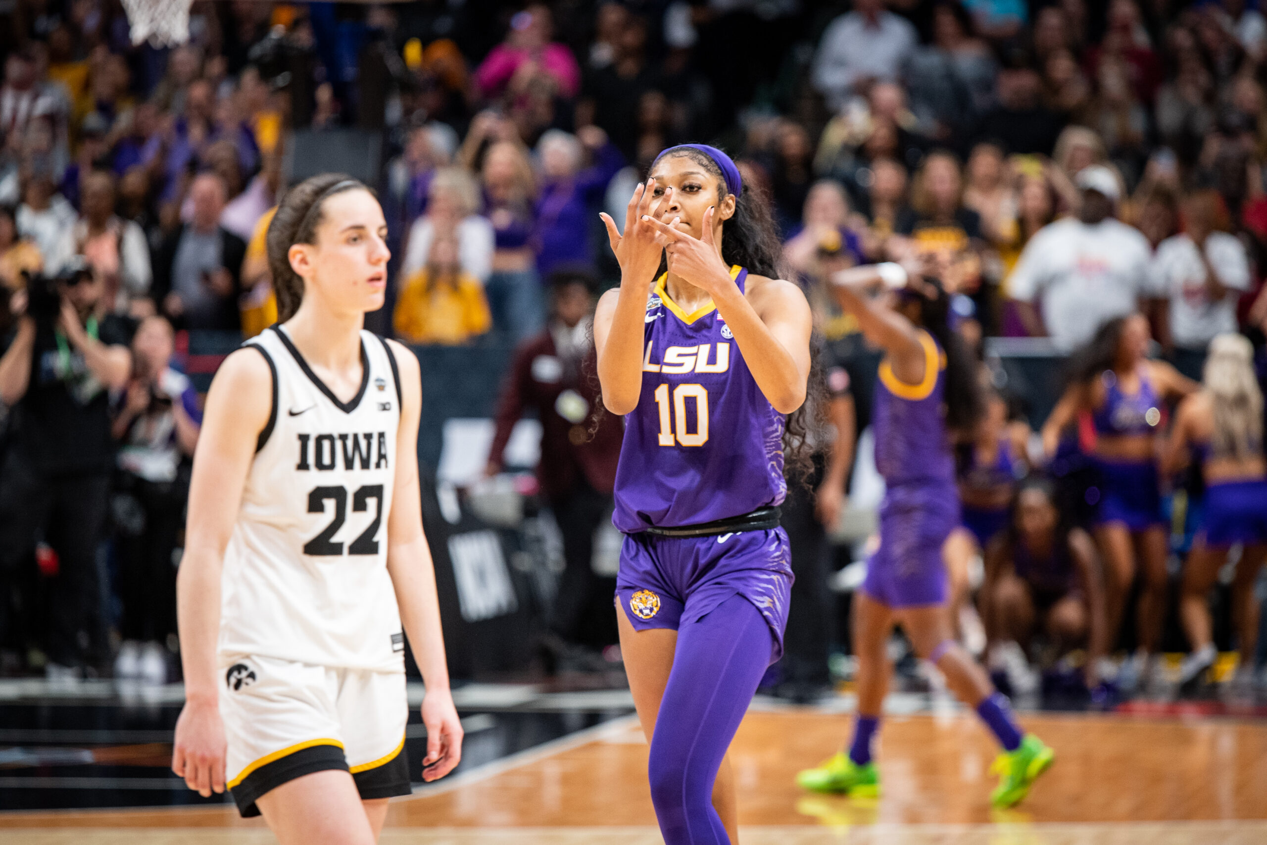 LSU forward Angel Reese points to her ring finger while celebrating a national title victory over Iowa on April 2, 2023. She is wearing purple and gold LSU away uniform. Opponent Caitlin Clark is in the forefront and wearing Iowa white and black home jersey.