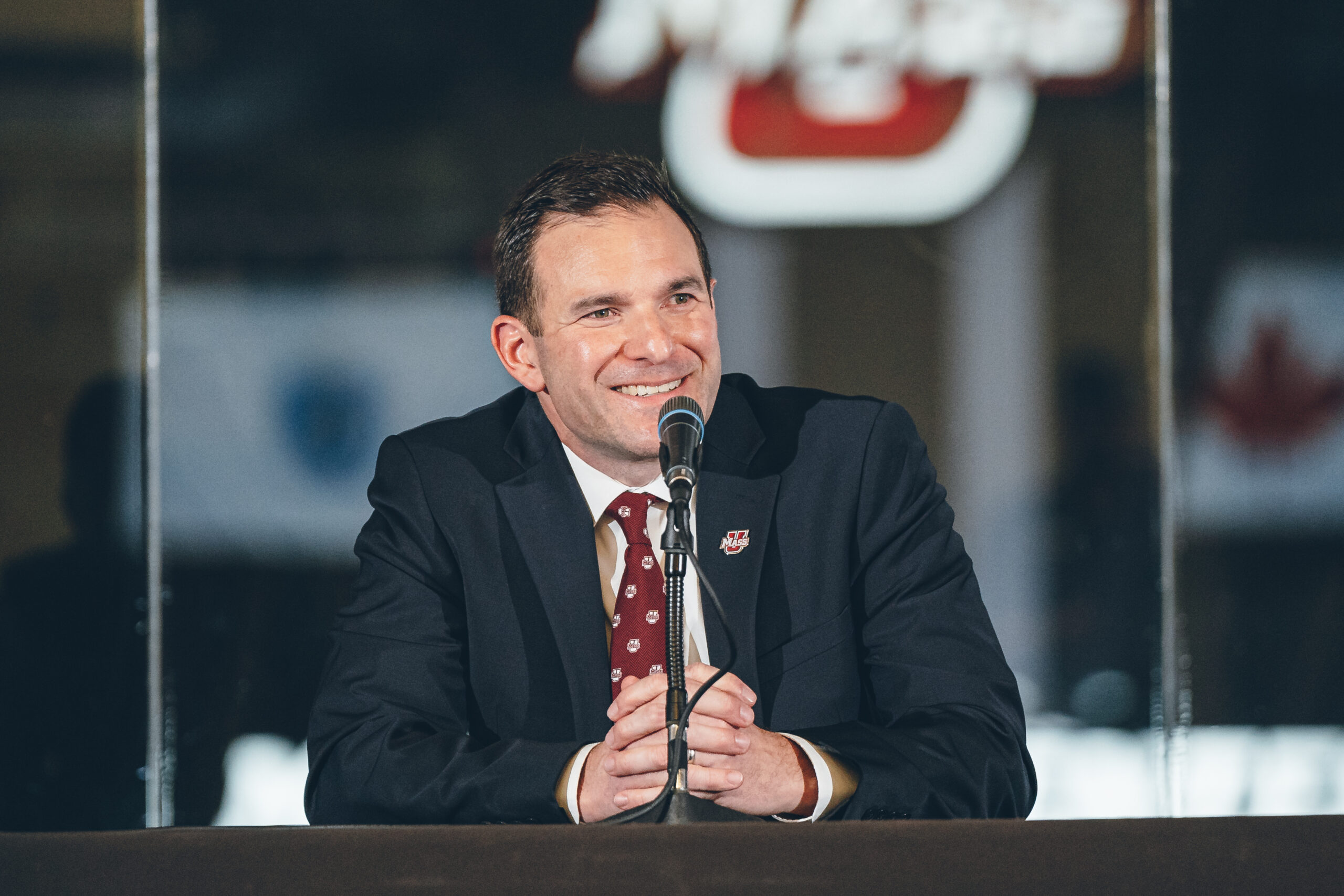 UMass head coach Mike Leflar smiles at his introductory press conference wearing a suit and UMass tie.