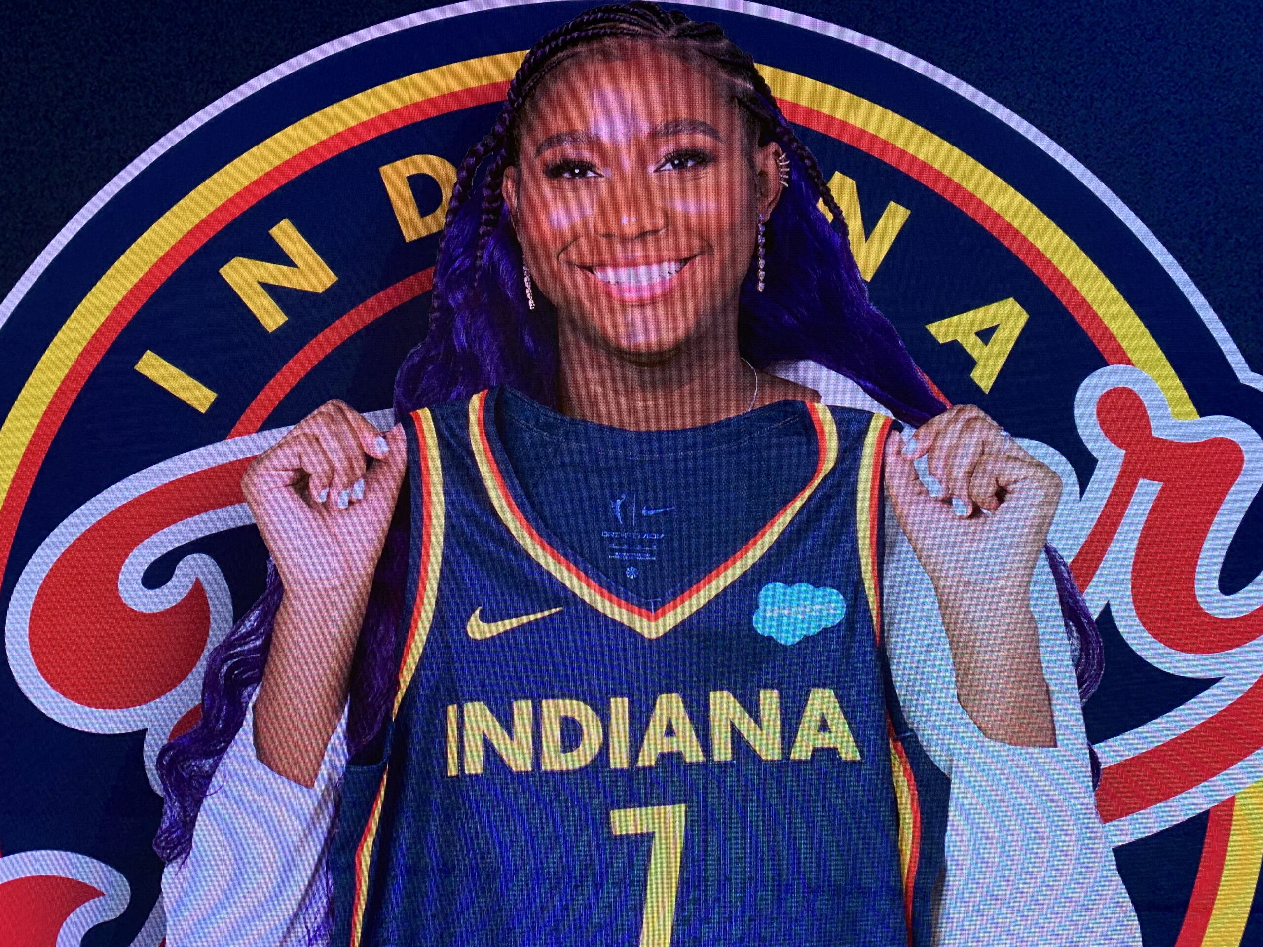 A sign inside Gainbridge Fieldhouse in Indianapolis shows Indiana Fever rookie center Aliyah Boston smiling and holding her blue No. 7 jersey at chest level. The Fever logo is behind her.