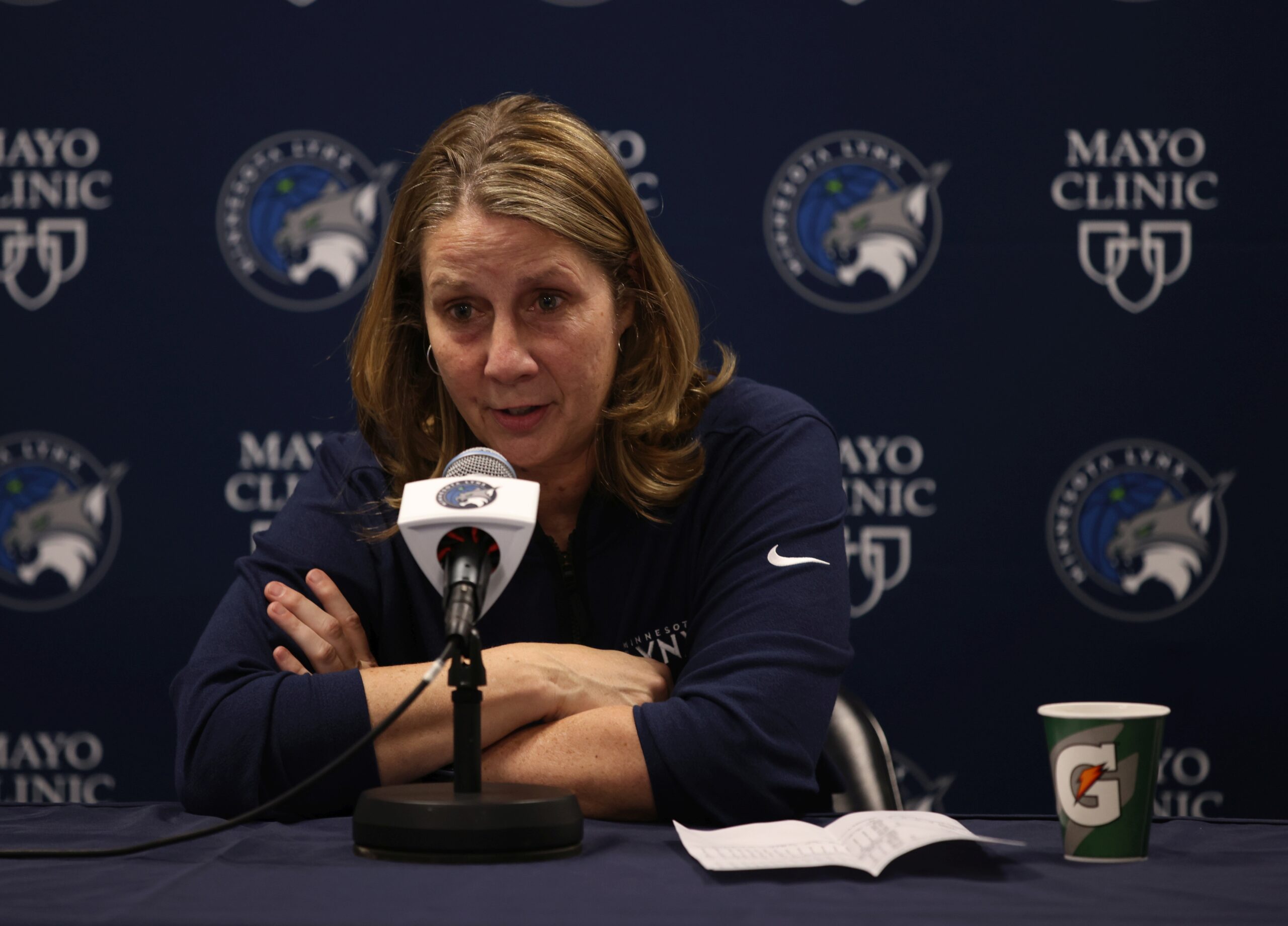 Minnesota Lynx head coach and president of basketball operations Cheryl Reeve sits at the podium and addresses the media. Her arms are crossed in front of her and resting on the table, and a backdrop behind her features Lynx and Mayo Clinic logos.