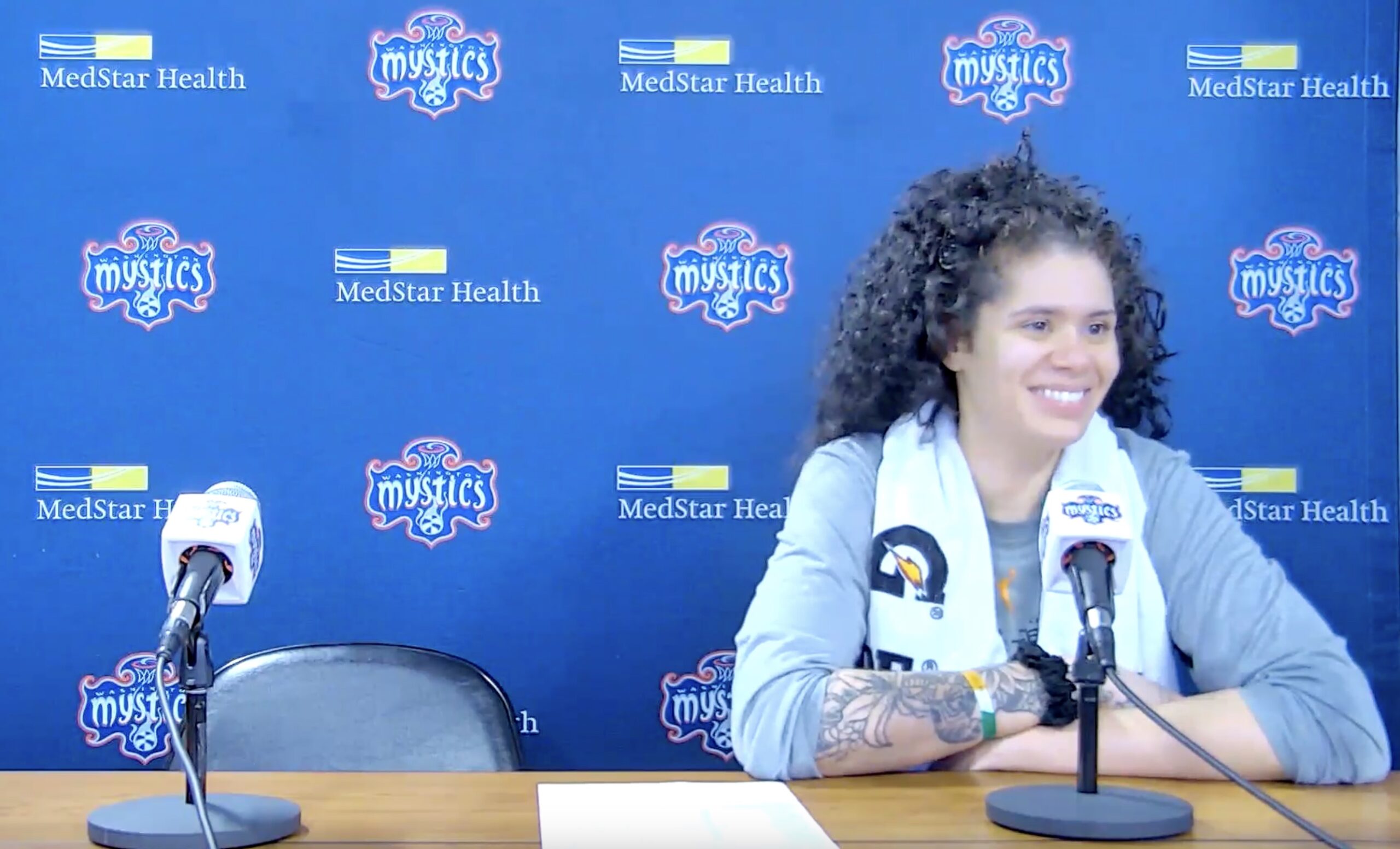 Washington Mystics center Amanda Zahui B. smiles as she sits at the podium talking to reporters. A backdrop dotted with Mystics and MedStar Health logos is behind her.