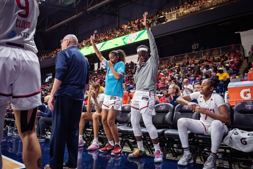 Standing in front of her seat on the bench, Washington Mystics guard Jazmine Jones puts her right hand to her face and makes a "three" sign with her left hand in celebration.