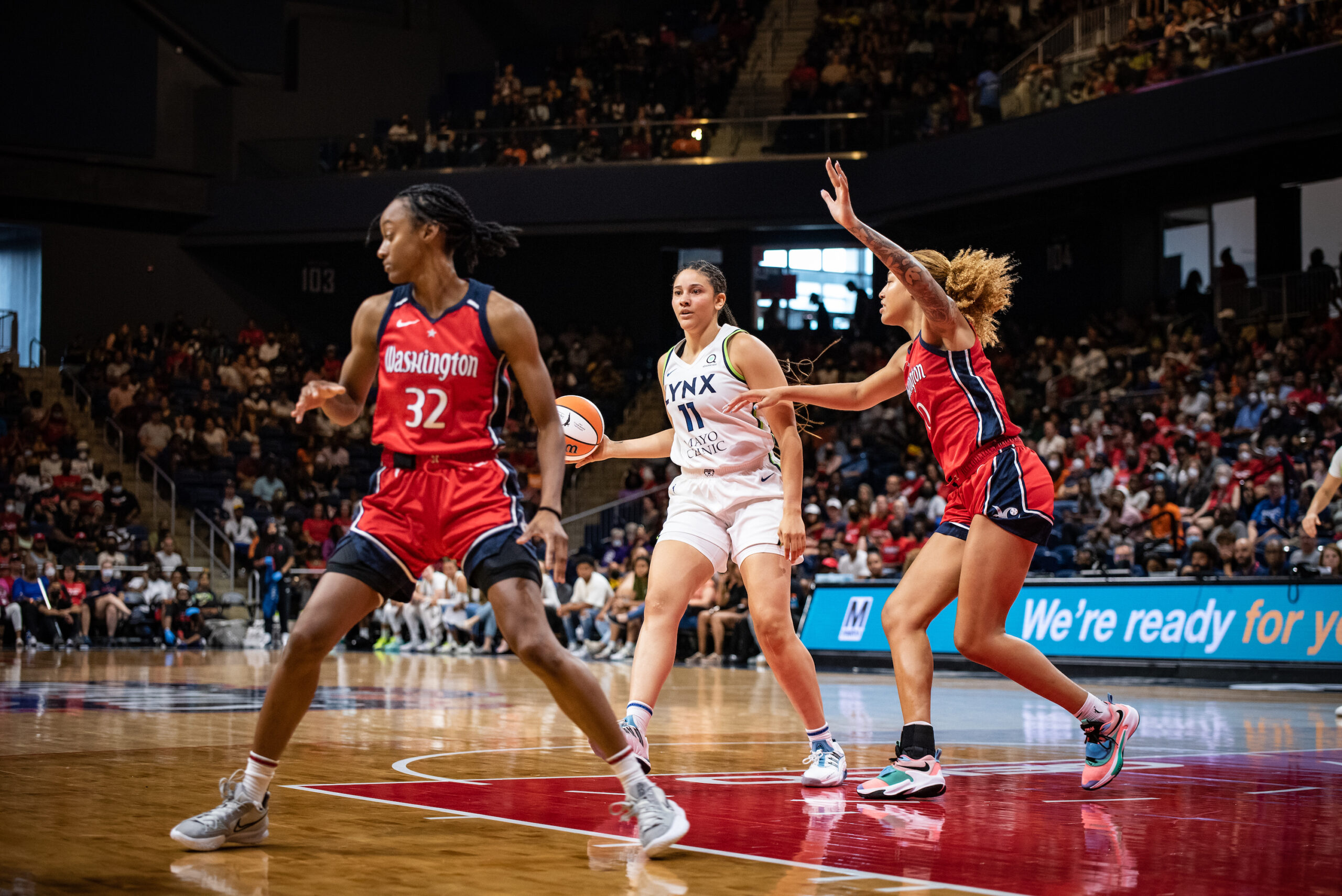 Minnesota Lynx forward Natalie Achonwa holds the ball and looks for a handoff near the elbow as Washington Mystics center/forward Shakira Austin defends. Mystics guard Shatori Walker-Kimbrough guards an off-camera Lynx player near the corner.