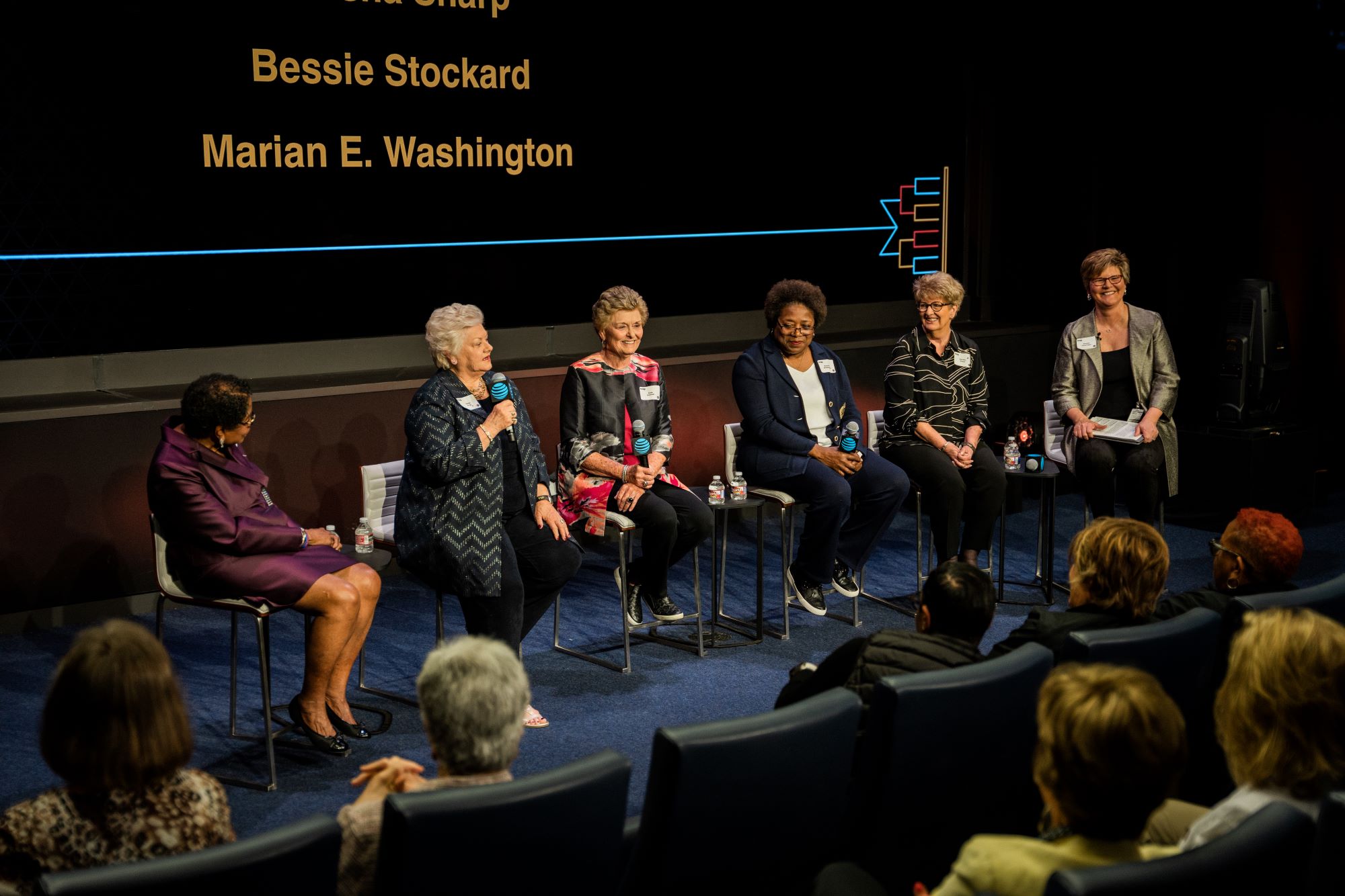 Six women sit on stage. The woman second from the left holds a microphone in her hand while answering the question she was asked.