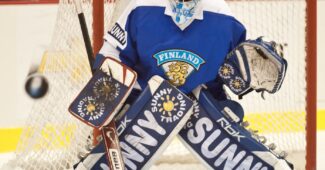 Goalie Noora Räty is positioned in front of the net wearing Finnish hockey gear.