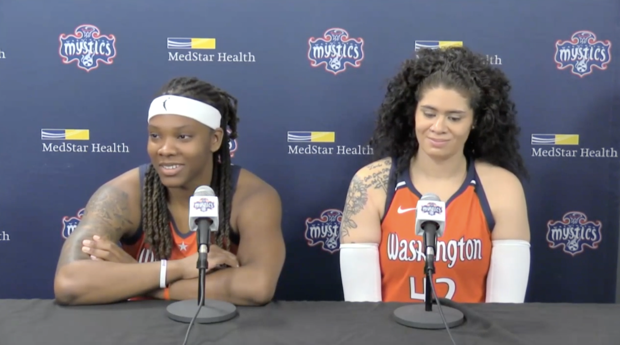 Washington Mystics forward Myisha Hines-Allen rests her arms on the table as she answers a reporter's question during a press conference. Center Amanda Zahui B. is on her left looking down at the table with a small smile.