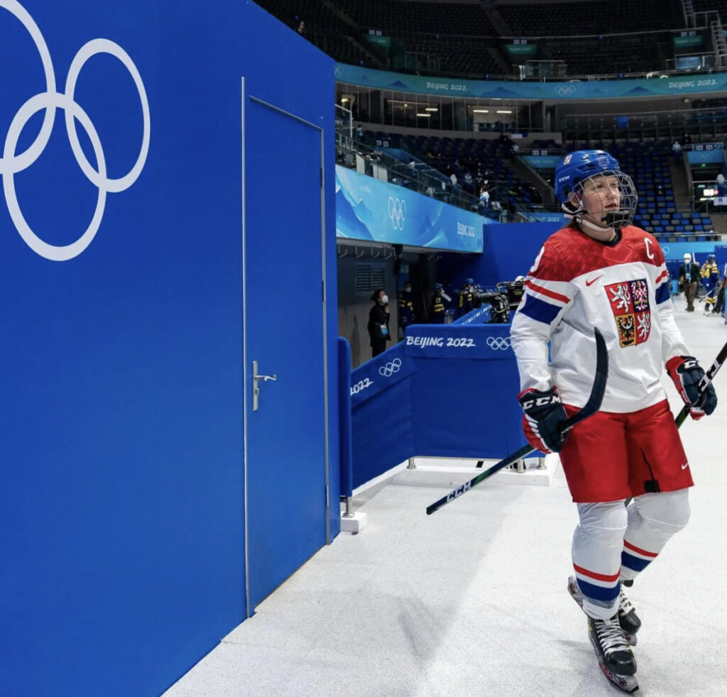 Czechia captain Alena Mills skates on the ice with Olympics symbol in the backdrop.