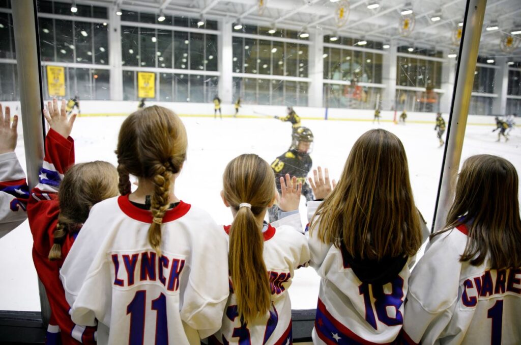 Young fans wearing Boston Pride jerseys watch from behind the glass as a game is in play on the ice.