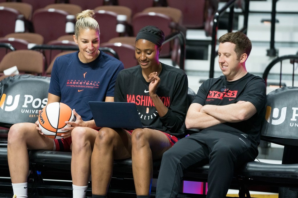 Washington Mystics forward Elena Delle Donne, associate head coach LaToya Sanders and head coach Eric Thibault sit on the bench together. Delle Donne is holding a ball, and they smile as they look at something on a laptop that Sanders has on her lap.