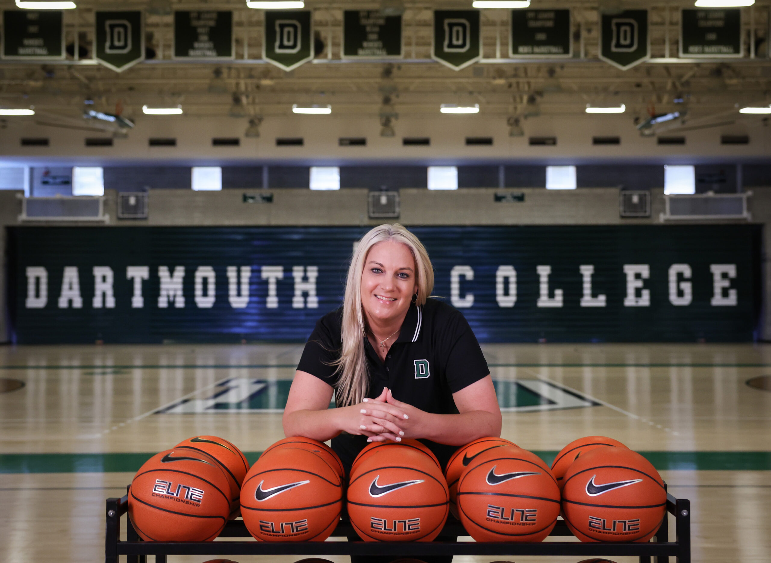 Dartmouth head coach Linda Cimino rests her elbows on a rack of basketballs and smiles for a photo.