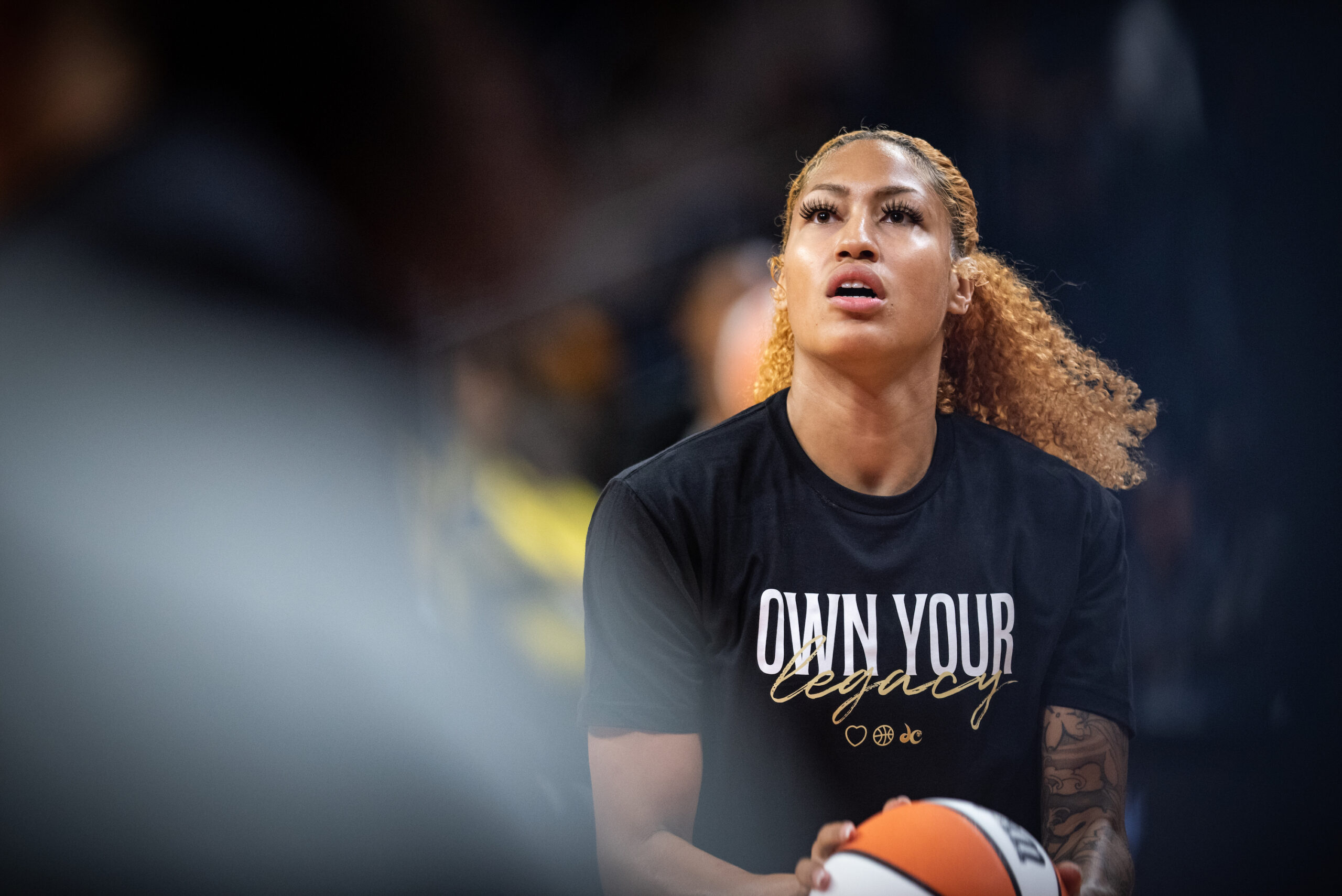 Washington Mystics center/forward Shakira Austin holds the ball in front of her at waist height as she warms up for a game. She is wearing a black warm-up shirt that reads, "Own your legacy."