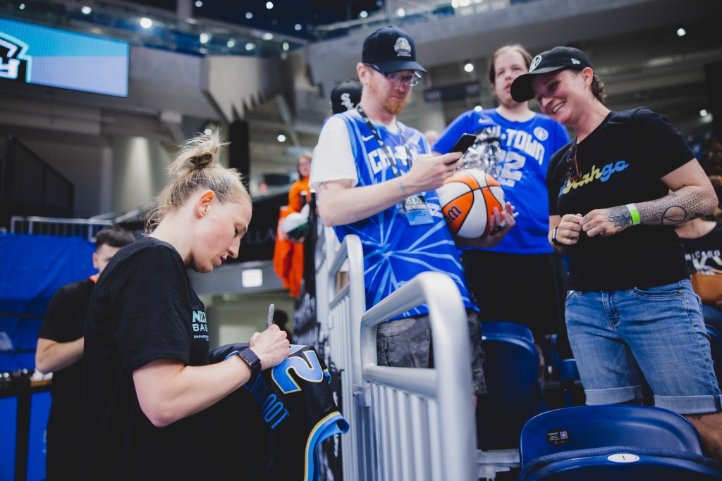 New York Liberty guard Courtney Vindersloon autographs a black Vandersloot Chicago Sky jersey as a Sky fan looks on and grins. Several fans wait in line to be next.