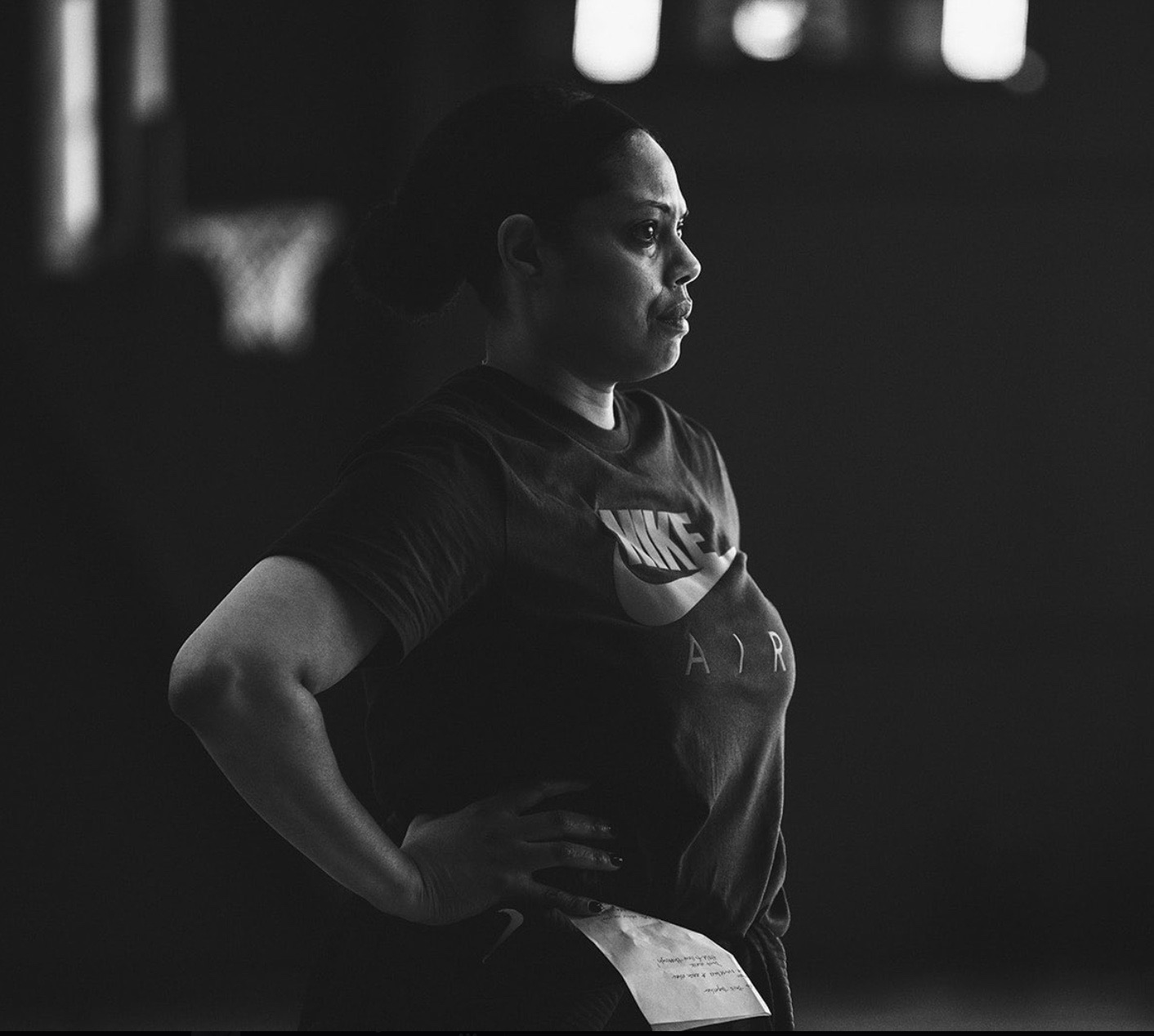 Xavier women's basketball head coach Billi Chambers is pictured in a black-and-white photo with her right arm on her waist and a basketball practice facility as the backdrop.