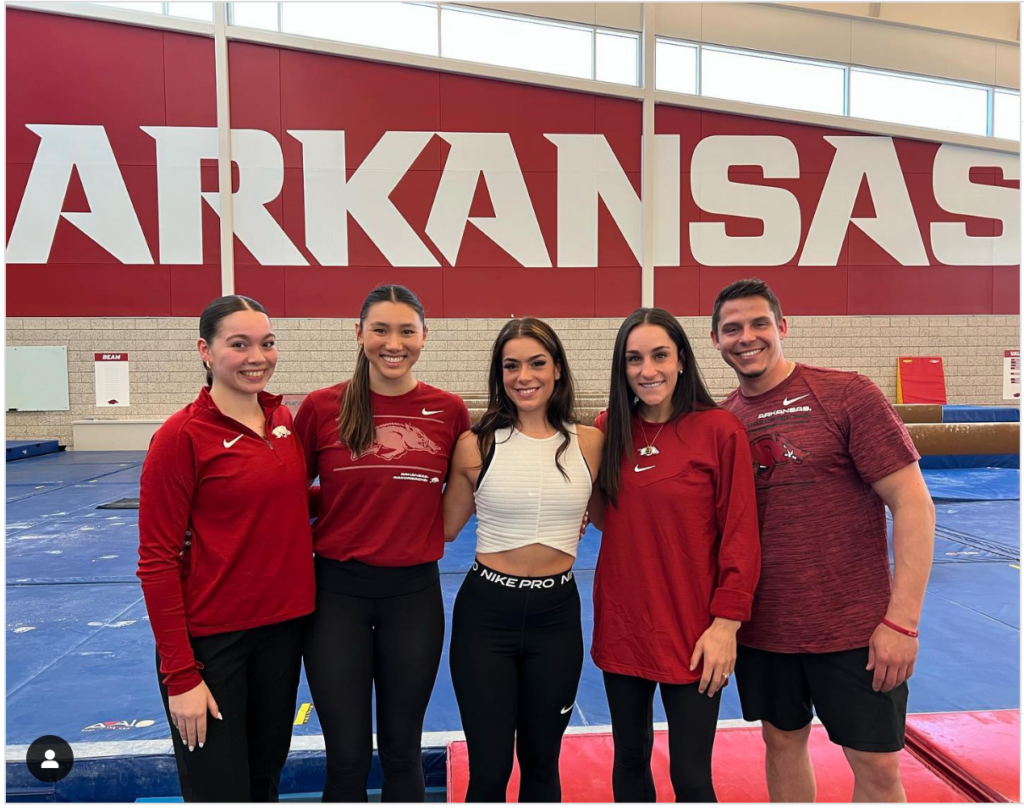 A posed photo in a gym of gymnast Sirena Linton with four Arkansas coaches, who are all wearing red Razorbacks gear. "Arkansas" is shown on the wall behind them in white lettering against a red backdrop.