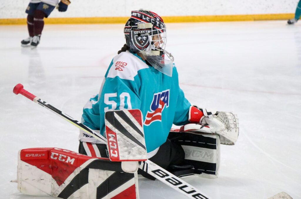 A goalie is pictured stretching on the ice wearing a blue USA Hockey jersey and participating in the USA Hockey - BioSteel Player Development camp.