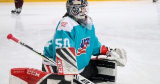 A goalie is pictured stretching on the ice wearing a blue USA Hockey jersey and participating in the USA Hockey - BioSteel Player Development camp.