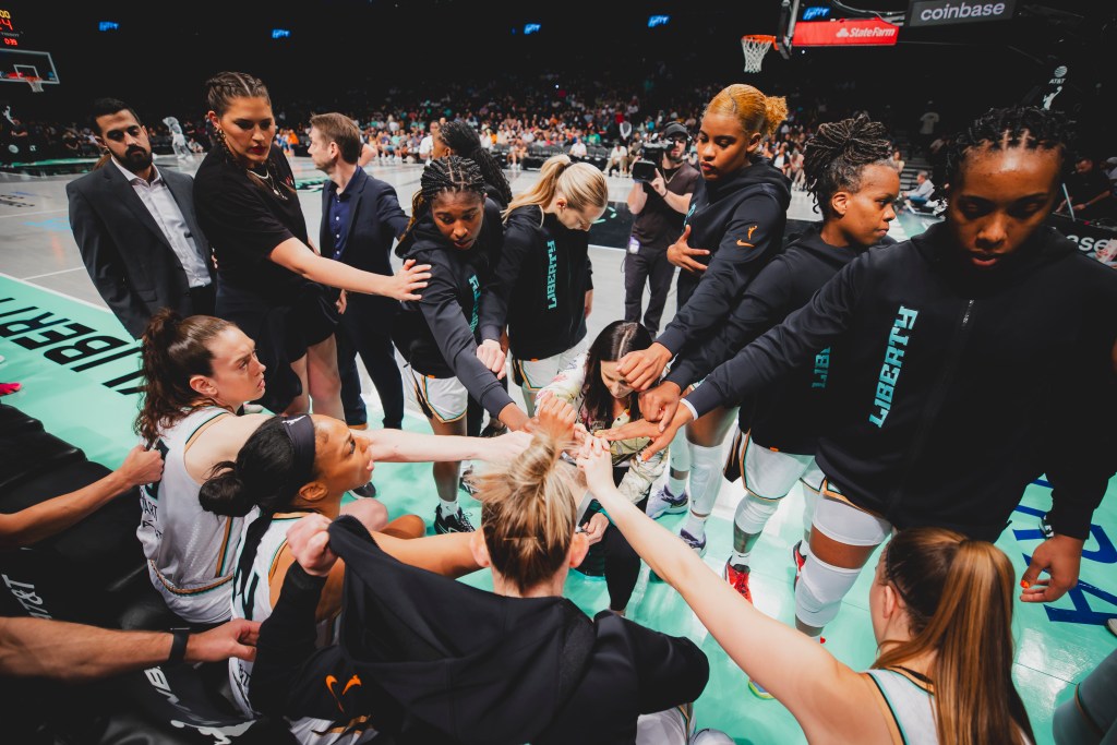The New York Liberty and their coaches all huddle with their hands in the middle during a timeout.