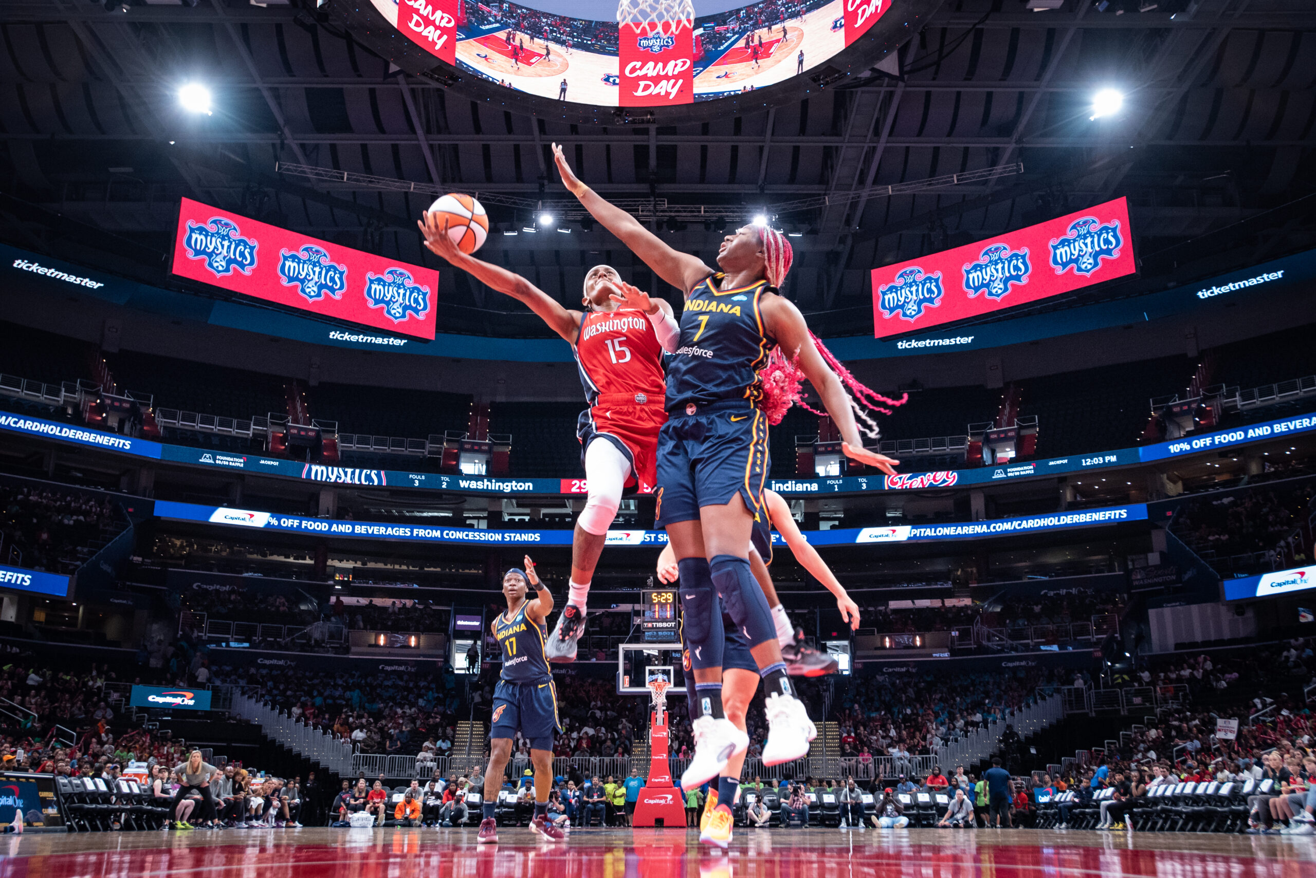 Washington Mystics wing Brittney Sykes holds the ball away from her body in midair while attempting a scoop layup around Indiana Fever center Aliyah Boston. Boston is meeting Sykes in midair with an arm outstretched toward the ball.