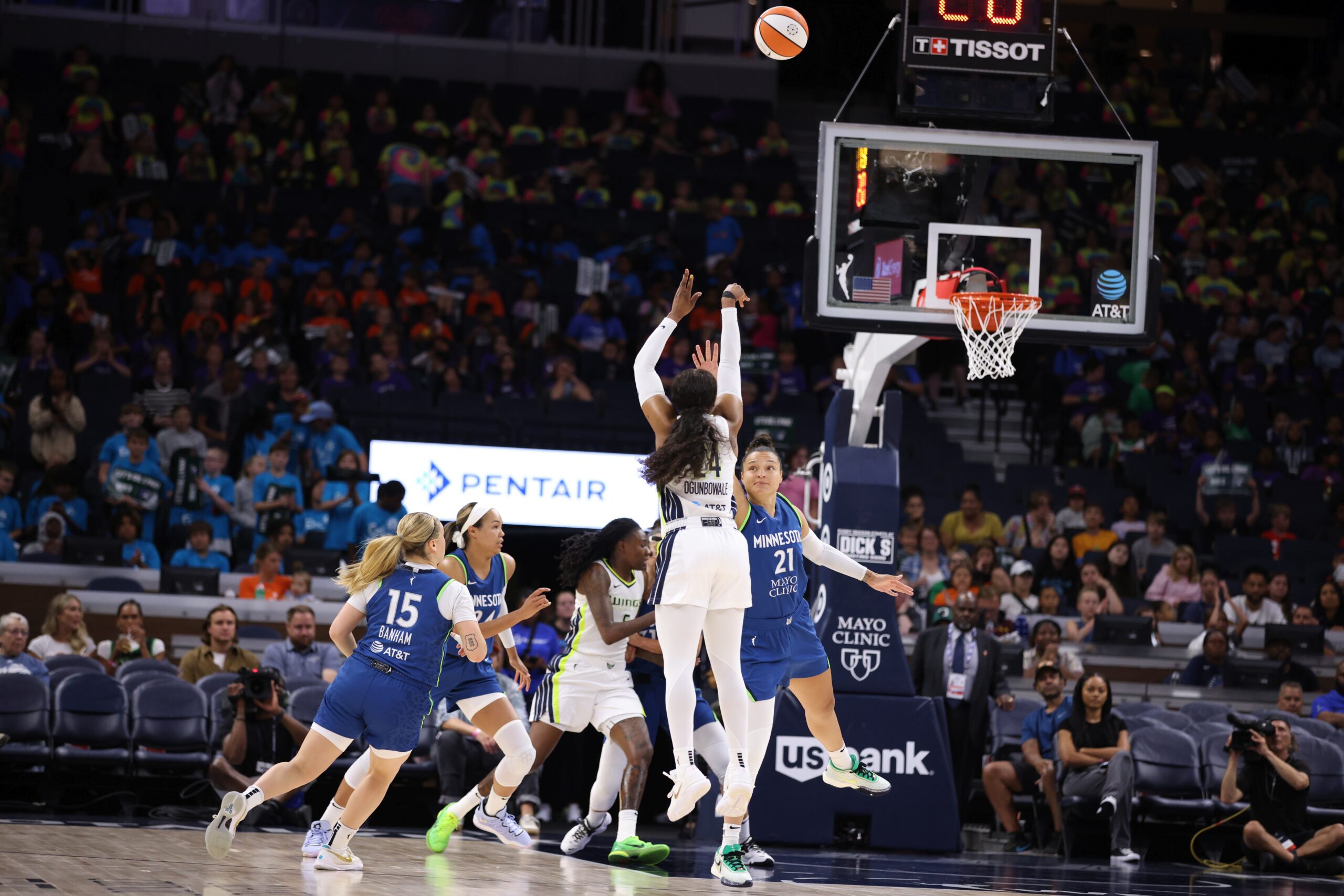 Dallas Wings guard Arike Ogunbowale holds her follow-through in midair, with a Minnesota Lynx defender lunging toward her, as the ball travels past her and toward the rim while players from both WNBA teams fight under the basket.