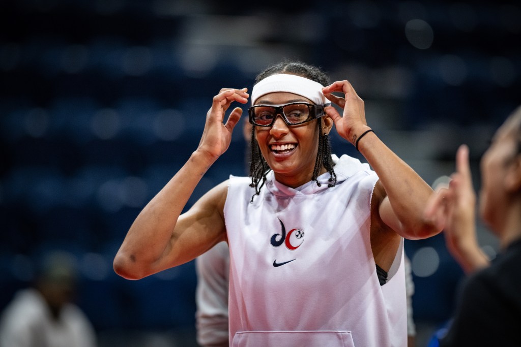 Washington Mystics guard Brittney Sykes is shown in a close-up photo. Wearing goggles and a headband, she smiles, and her hands are near her head, as if she's about to or has just adjusted her goggles or headband.