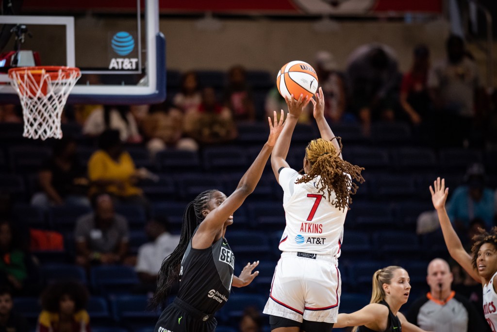 Washington Mystics guard Ariel Atkins is shown from behind. She is in the air about to release a 3-pointer as Seattle Storm center Ezi Magbegor stretches her right arm up to contest.