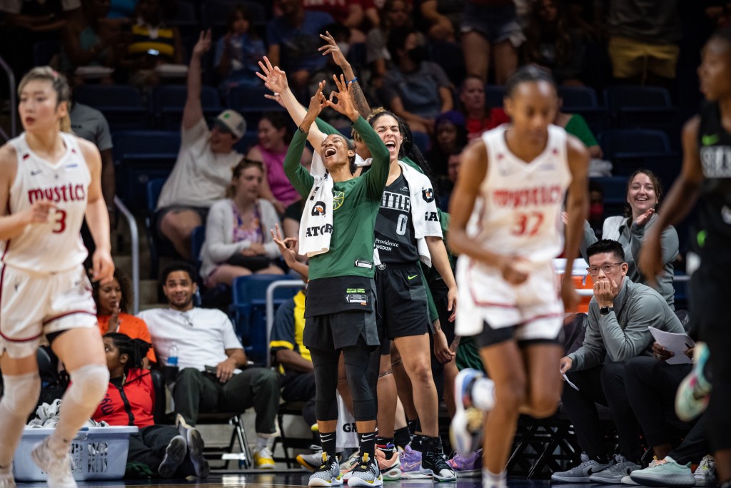 Three Seattle Storm players hold their hands up in celebration, some of them making the 3-point sign. Two Washington Mystics players run up the court in the background.