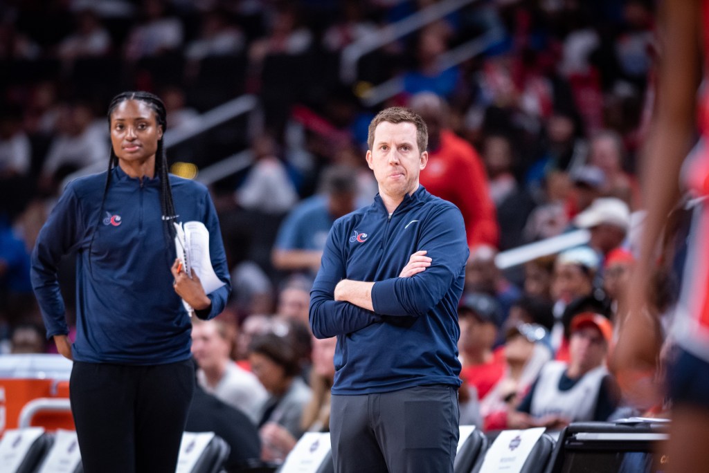 Washington Mystics head coach Eric Thibault stands on the sidelines with his arms folded across his chest. Associate head coach LaToya Sanders looks on from a few steps behind him.