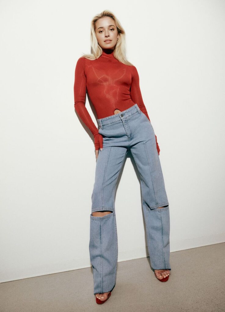 LSU gymnast Livvy Dunne is posing for a photograph while wearing a red, long-sleeved top and faded, high-waisted blue jeans.