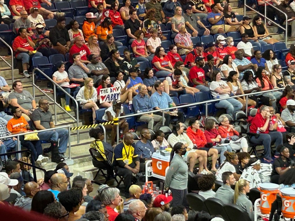 A wide photo of the crowd at a Washington Mystics game. One fan holds a sign reading, "The DMV โค๏ธ Abby," referencing Abby Meyers.